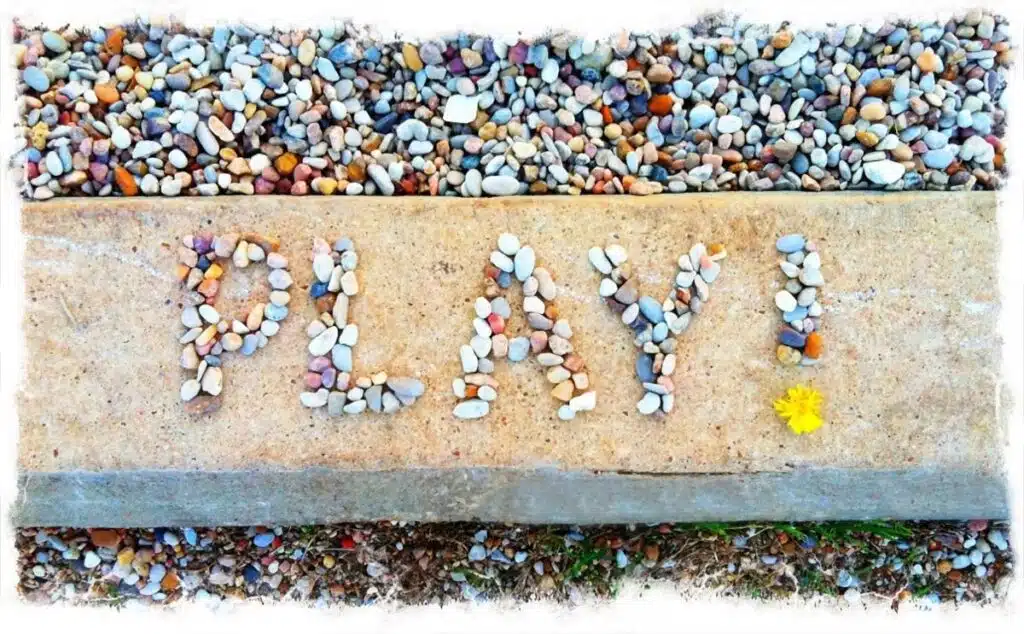 The word "play!" spelled out using small, colorful pebbles